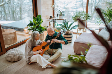 Senior couple playing on guitair, sitting in cozy living room and enjoying autumn day.