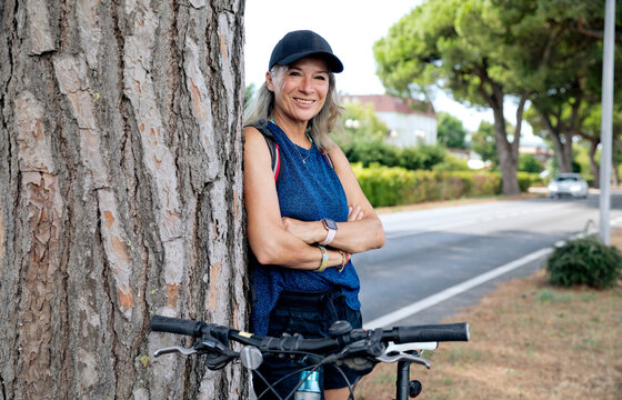 Smiling senior woman with arms crossed leaning on tree trunk by bicycle