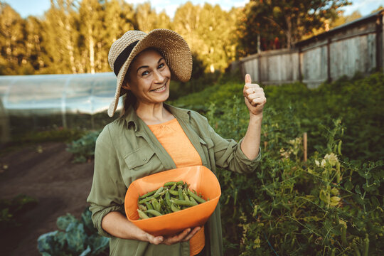 Smiling woman with bowl of green peas gesturing thumbs up in garden - Powered by Adobe
