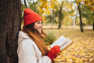 Happy young woman holding book by tree trunk at park