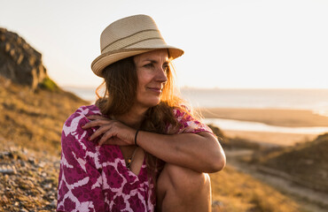 Smiling mature woman wearing hat sitting on beach at sunset