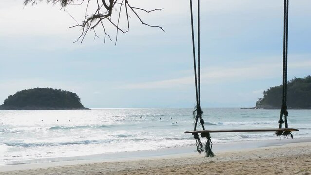 Wooden swing on the sandy beach, behind them are people play surfboard  sea at the evening. with blue sky and the turquoise  sea. Kata beach Phuket Thailand. Concept of summer vacation, relax.