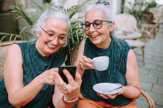 Happy senior women twins having coffee break in city, smiling and checking phone.