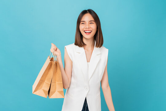 Cheerful beautiful Asian woman in a white t-shirt and holding brown blank craft paper bag on blue background.
