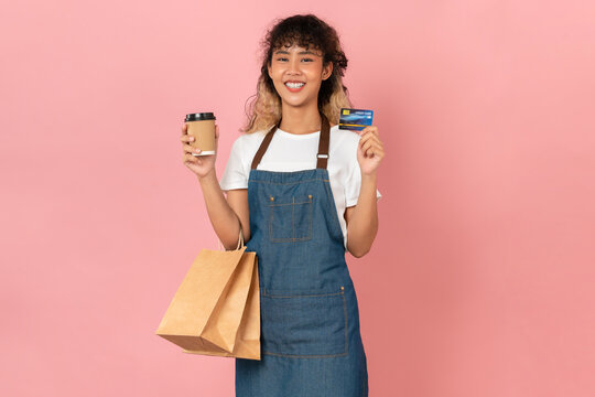 Happy young Asian woman wears an apron and holding brown blank paper shopping bags and showing credit card on pink background.