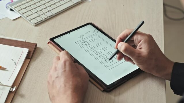 Closeup of unrecognizable app developer using digital tablet while drawing scheme for his new development, working at desk in office