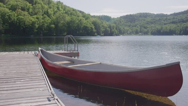 Static shot of red canoe floating in calm lake water next to wooden dock in forested lake in Laurentian Mountains in Quebec Canada