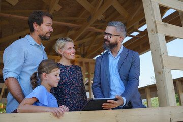 Sales agent showing plans of new unfinished ecologic wooden house to young family on construction...