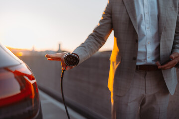 Businessman, holding power supply and charging his electric car during sunset. Concept of ecology...