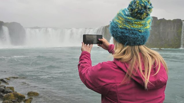 Female hiker traveler in the rain photographs godafoss  waterfall iceland,woman standing in front of cascade taking photo picture using smart phone mobile