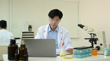 Young male Biochemist using laptop computer, collecting data and summarising information for scientific report
