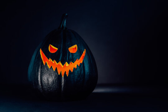 Spooky Halloween Black Pumpkin, Jack O Lantern, With Orange Illuminated Evil Face And Eyes On Black Background With Copy Space. Happy Halloween Decor Concept. Festive Postcard. Selective Focus