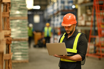 Warehouse manager wearing hardhats and reflective jackets using laptop for checking stock and order details in warehouse