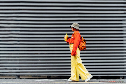 Stylish girl in bright clothes drinking sugar flavored tapioca bubble tea while walking near gray striped urban wall. Full length portrait of fashionable hipster girl. Street fashion. Copy space.