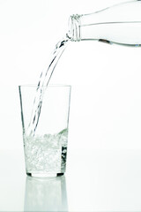 Detail of a glass bottle pouring water into a glass tumbler on a white background.