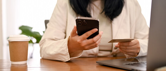 A female holding a smartphone and a credit card while sitting at her office desk. cropped shot