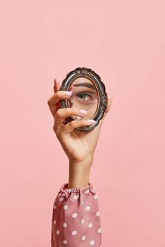 Retro style. Female hand holding small mirror with reflection of girl's eye isolated over pink background. Concept of style, beauty, art