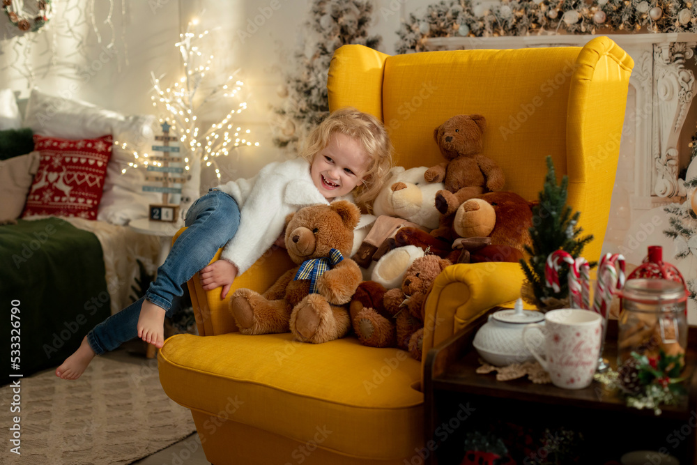 Wall mural Cute little toddler curly girl in winter christmas sweater, sitting in cozy room, decorated for christmas