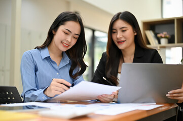 Cheerful two Asian young businesswomen working together on sales and financial report.