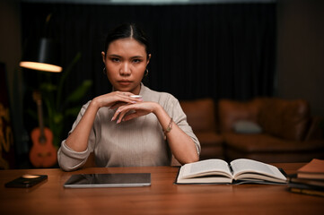 Confident Asian businesswoman looking at the camera, sitting at her office desk at night.