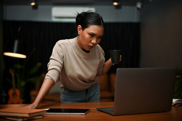 Obraz premium Confident Asian businesswoman leaning on table, looking at her laptop screen, sipping coffee