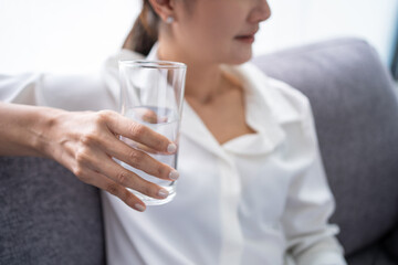 Young asian woman holding glass of pure mineral aqua water in hands and sitting on comfortable sofa at home, feeling refreshed energetic in morning.