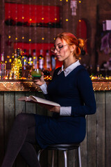 A red-haired girl is sitting in a bar with a book in her hands
