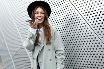 a young woman entrepreneur in an autumn coat and a black hat speaks on a mobile phone with a speakerphone against the background of an aluminum facade of a business center - obrazy, fototapety, plakaty