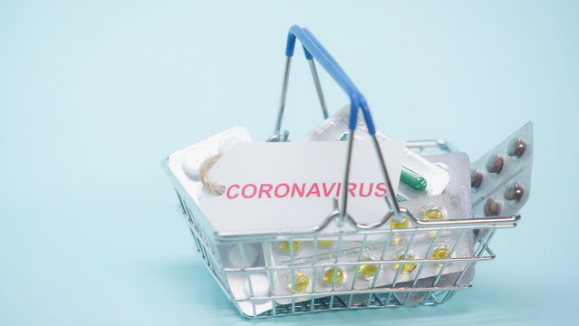 Shopping Basket With Coronavirus Lettering On Card And Pills On Blue.