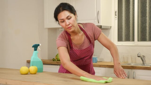 Asian woman in apron washing top of kitchen table during cleanup in apartment.