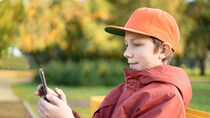 A teenage boy sits on a wooden bench in the park in autumn and uses a mobile phone, watches videos, surfs the Internet. Video games, social media addiction.