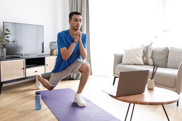 Obraz premium Young arab guy doing leg exercises on yoga mat in living room interior, watching online workout on laptop