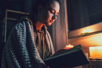 Teenage girl sits under blanket near heating radiator with candles and read book .Rising costs in private households for gas bill due to inflation and war, Energy crisis