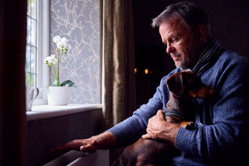 Mature Man With Pet Dog Trying To Keep Warm By Radiator At Home During Cost Of Living Energy Crisis