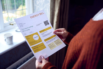 Naklejka premium Senior Woman With Euro Energy Bill Trying To Keep Warm By Radiator In Cost Of Living Energy Crisis