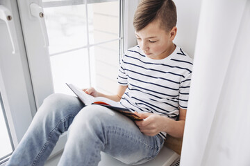 Portrait of a handsome teenager reading a book, sitting on windowsill in a cozy home.