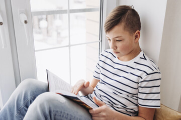 Portrait of a handsome teenager reading a book, sitting on windowsill in a cozy home.