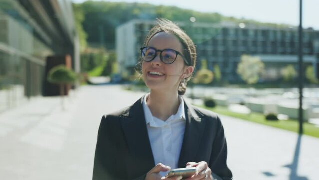 Footage of smiling cute young Caucasian woman with elegant hairstyle holding smartphone, chatting, using navigator, looking for adress sign. Meeting during lunch time. Blurred background of modern