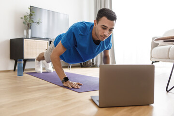 Handsome arab guy in sportswear doing push-ups, having online fitness class via laptop from home, copy space