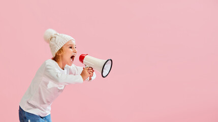 Studio shot of little girl, kid in casual style clothes and warm hat shouting at megaphone isolated...