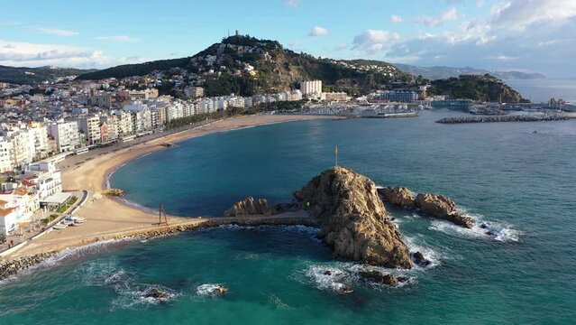 Scenic drone view of coastal Catalan town of Blanes overlooking white residential buildings and Sa Palomera Rock on background of San Juan hill with ancient castle on sunny day, Barcelona, Spain 