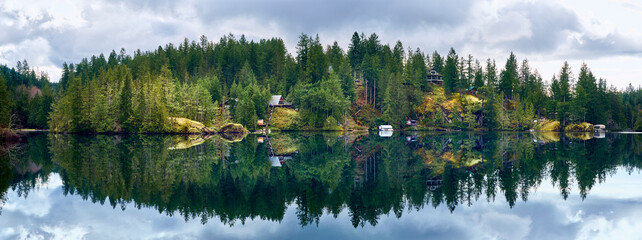 Houses on the rocks among the coniferous forest are reflected in the crystalline water of the wild Ruby Lake in calm weather. Forest landscape at Dan Bosch Park, Sunshine Coast Hwy, BC, Canada
