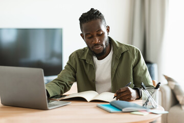 Obraz premium Focused african american man sitting at desk working on laptop and taking notes in notebook, studying online at home