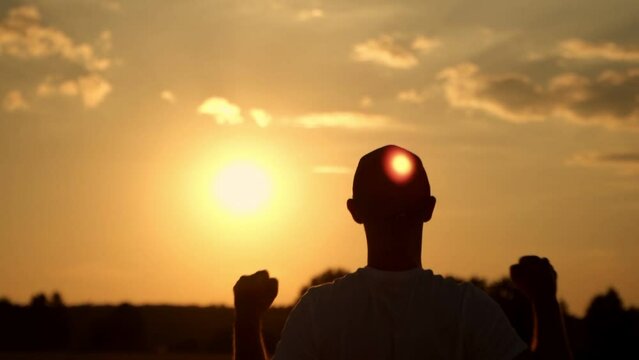 The silhouette of a man at sunset stands with his hands raised with happiness