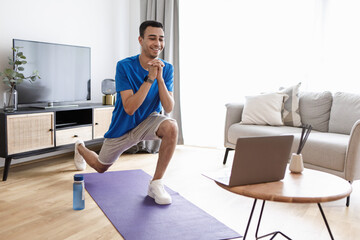 Happy arab man doing forward lunge exercises while watching online workout tutorial via laptop,...