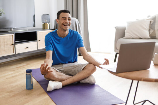 Positive calm arab man in sportswear sitting in lotus pose, having online yoga class, meditating near laptop at home