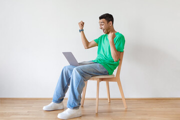 Overjoyed arab man using laptop, shaking fists and shouting while sitting on chair over light wall,...