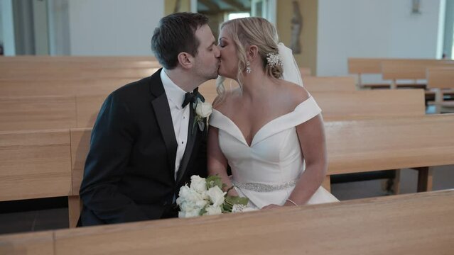 Bride and groom kissing while sitting on a wooden pew in a modern church. Dolly shot.
