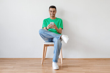 Excited young arab man using cellphone and texting or networking in social media sitting on chair over light wall