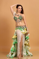 a pretty girl brunette who dances go-go and belly dance, stretching posing in studio in green arabian dress on a beige background